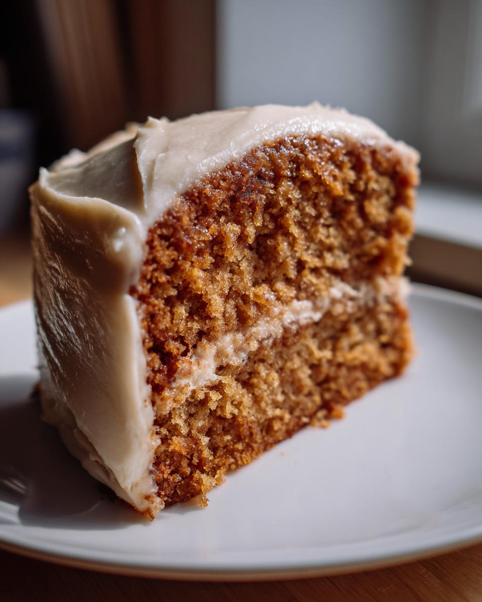 Close-up of a moist slice of applesauce cake layered with light frosting, sitting on a white plate.