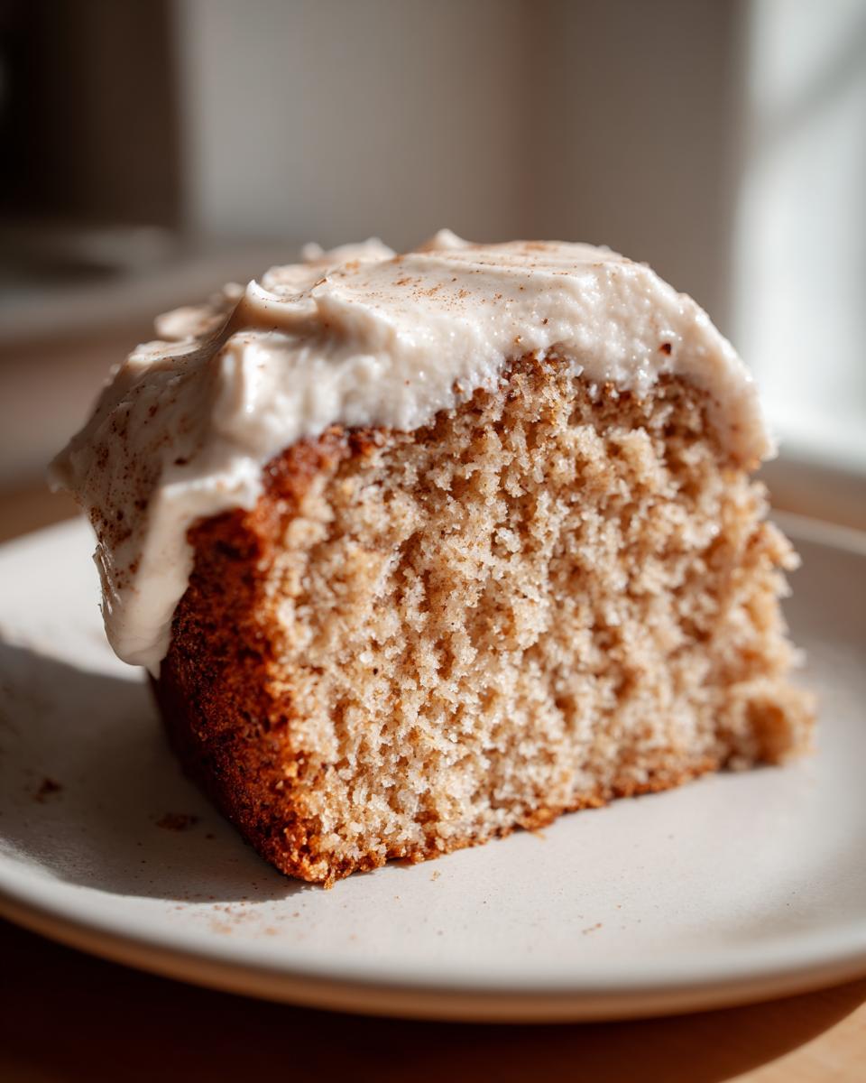 Close-up of a moist slice of applesauce cake topped with thick, creamy frosting dusted with cinnamon.