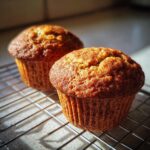 Two freshly baked, golden brown moist bran muffins cooling on a wire rack in bright sunlight.