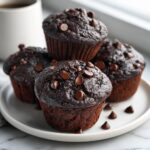 A stack of four rich, dark chocolate zucchini muffins topped with chocolate chips on a white plate.