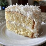 Close-up of a moist slice of coconut cake recipe, layered with white frosting and topped with shredded coconut flakes.