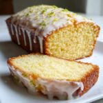 A slice of moist lemon pound cake sits in front of the loaf, both covered in a thick white glaze and bright green lemon zest.