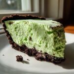 A thick slice of bright green, creamy no-bake grasshopper pie with a dark chocolate cookie crust, sitting on a white plate.