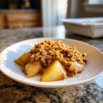 A close-up of a serving of warm pear crisp, featuring caramelized pear slices under a thick, crunchy oat crumble topping.