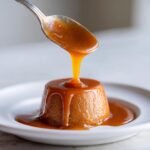 A spoonful of rich, glossy salted caramel sauce being poured over a small, round dessert on a white plate.