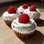Four delicious mini cheesecake treats topped with creamy frosting and a fresh raspberry, sitting on a wooden surface.