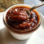 A spoonful of thick, dark homemade apple butter being lifted from a small glass jar.