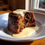 A rich, dark plum pudding with a slice taken out, topped and surrounded by creamy yellow custard.