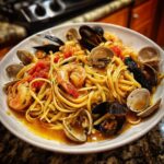 A close-up, appetizing view of a bowl of seafood pasta featuring linguine, shrimp, mussels, and clams in a rich tomato broth.