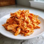 A mound of bright orange, shredded buffalo chicken piled high on a white plate.
