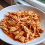 Close-up of shredded chicken tinga coated in a rich, red, spicy sauce, served in a white bowl.
