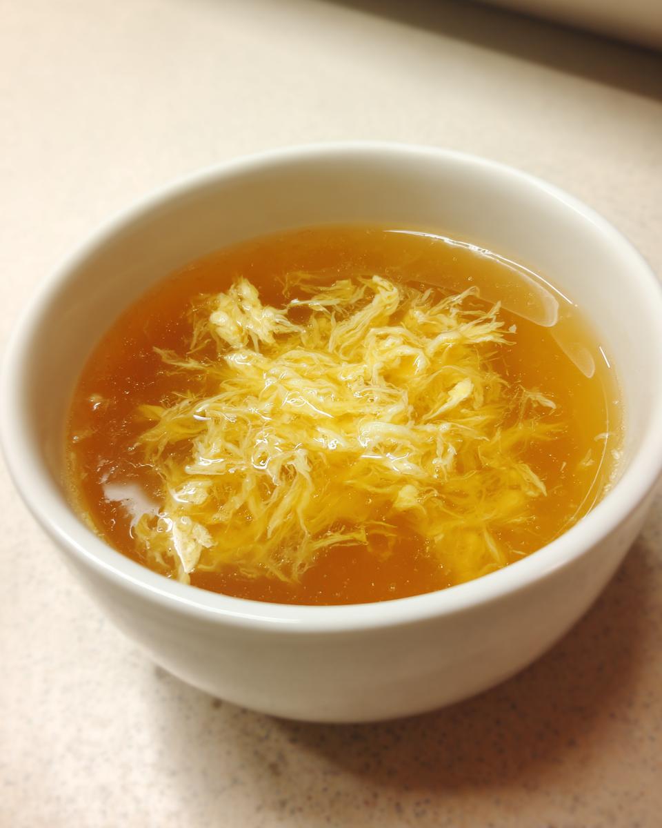 Close-up of a white bowl filled with golden broth and delicate strands of egg drop soup.