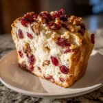 A close-up of a thick slice of fluffy cranberry walnut bread, generously topped with dried cranberries and walnuts.