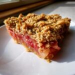 A close-up, sunlit slice of rhubarb crisp showing bright pink filling and golden oat topping on a white plate.