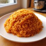 A perfectly mounded serving of bright orange, smoky jollof rice on a white plate.