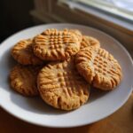A stack of soft peanut butter cookies with classic fork crosshatch marks resting on a white plate near a window.