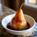 A single, beautifully glazed poached pear served upright in a small white bowl with dark syrup.