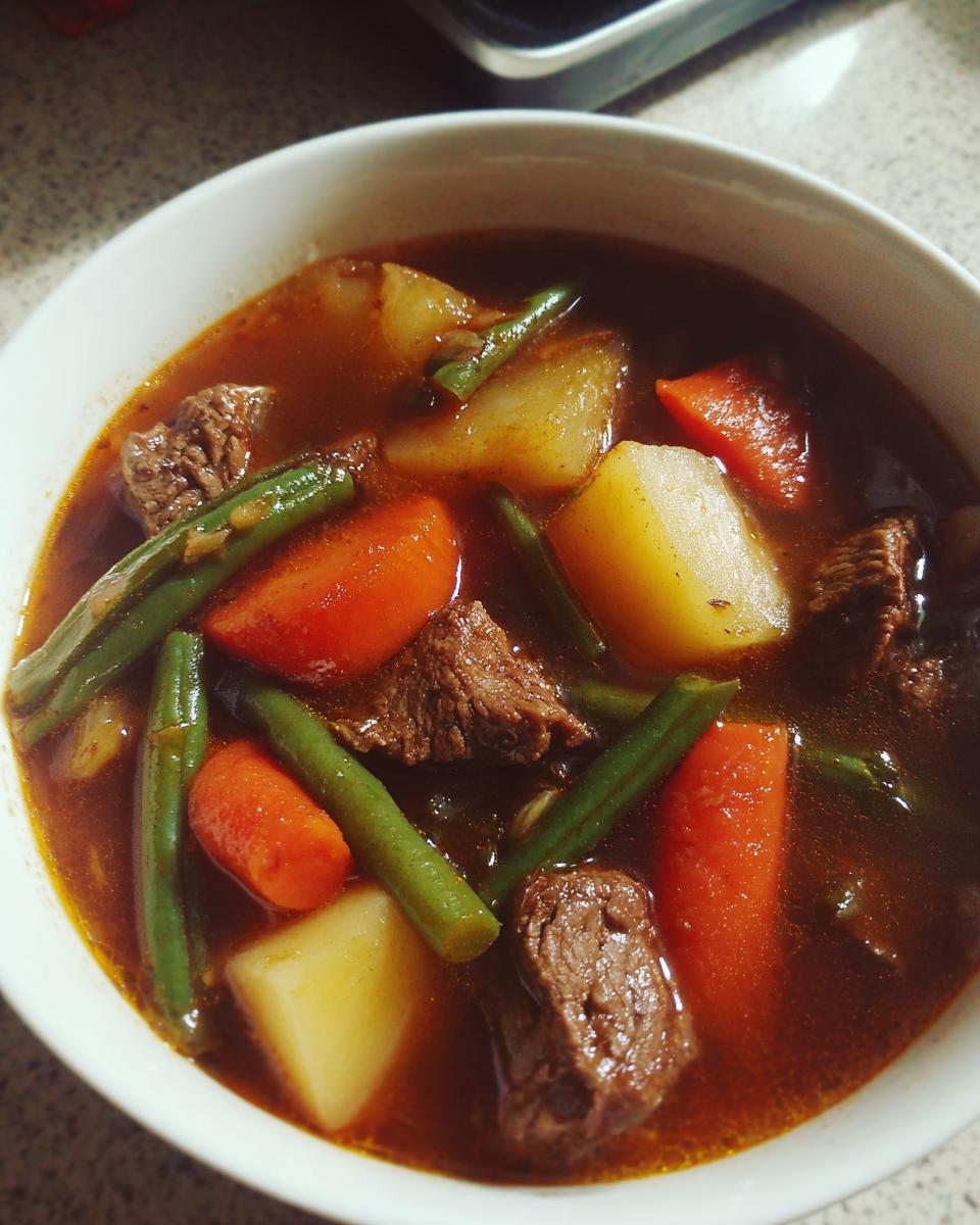 Close-up of a white bowl filled with rich vegetable beef soup, showing chunks of beef, potatoes, carrots, and green beans.