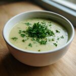 A close-up of a bowl of creamy, velvety celery soup topped with a generous mound of fresh chopped parsley.