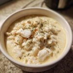 Close-up of a white bowl filled with rich, creamy crab soup, generously topped with large flakes of white crab meat and dried herbs.