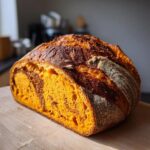A freshly baked, deeply colored pumpkin sourdough loaf with a striking orange interior, resting on a wooden cutting board.
