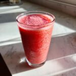 A tall glass filled with a bright red, icy watermelon smoothie sitting on a sunlit white marble counter.