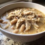 A close-up of a bowl filled with creamy white chicken chili, featuring shredded white chicken and cannellini beans, topped with cheese.