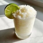 A frosty white Christmas margarita topped with shredded coconut and garnished with a fresh lime wheel.