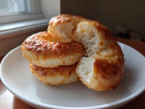 A stack of golden brown, cheesy high protein sourdough discard bagels on a white plate, one broken open showing the fluffy interior.
