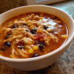 Close-up of a white bowl filled with creamy chicken enchilada soup crock pot, showing shredded chicken, black beans, and corn.