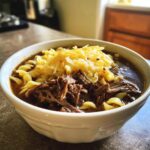 A close-up of a white bowl filled with crockpot french onion soup beef and noodles, topped with melted cheese.