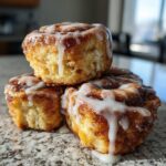 Three gooey gluten free sourdough discard cinnamon rolls stacked and dripping with white icing on a granite counter.