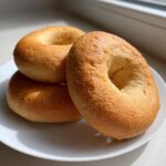 Three golden-brown high protein sourdough discard bagels stacked on a white plate near a window.