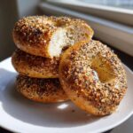 A stack of three everything sourdough discard bagels, with the top one cut in half showing the soft interior.