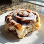 A close-up of a fluffy sourdough discard cinnamon roll generously topped with white icing, sitting on a white plate.