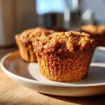 A close-up of a freshly baked peanut butter banana bread muffin topped with a crumbly streusel, sitting on a light plate.