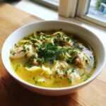A close-up of a white bowl filled with savory chicken penicillin soup, featuring chunks of chicken, egg noodles, and topped generously with fresh dill.