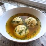 Close-up of a white bowl filled with golden broth and three fluffy matzo balls in this jewish penicillin soup, garnished with dill.