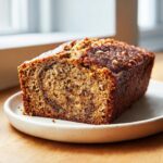 A freshly baked loaf of peanut butter banana bread, showing a moist crumb and dark crust, served on a small plate.