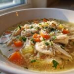 Close-up of a steaming bowl of penicillin soup featuring shredded chicken, carrots, orzo, and grated cheese.